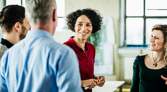 A group of colleagues smile while talking in a bright open office setting with windows in the background.