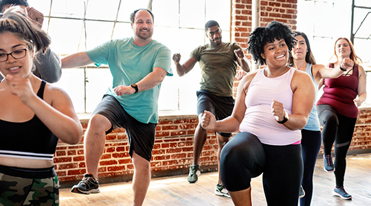 A group of men and women smile while doing aerobics in a bright studio with windows in the background.