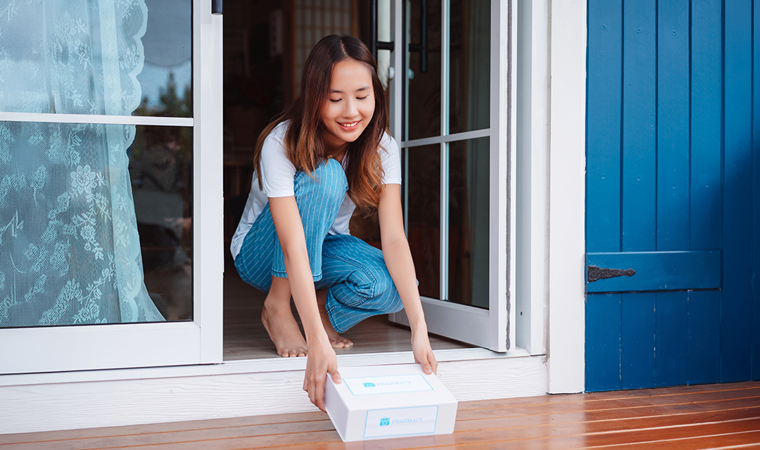 A young woman bends down to pick up a mail-order medication box at her doorstep.