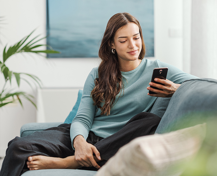 A young woman sits comfortably on a couch smiling as she looks at her phone in a comfortable home setting.
