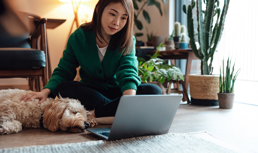 A woman sits on the floor petting her dog while checking her laptop.