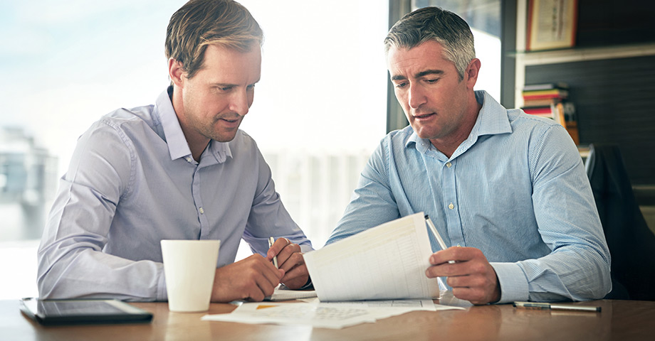Two well-dressed colleagues seated at an office table reviewing and discussing reports.