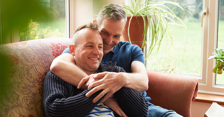 Male couple sit on couch hugging and laughing together.