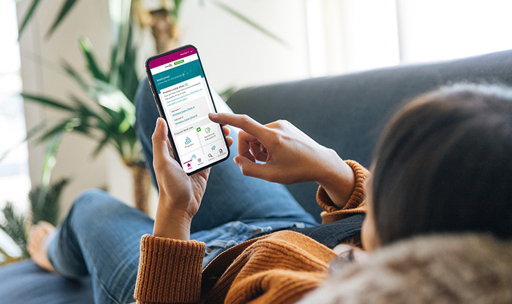 Young woman relaxes on couch, viewing Member Dashboard on mobile phone.