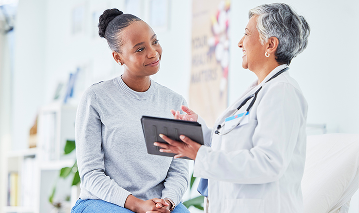 Elderly doctor holding mobile tablet smiles while female patient listens attentively.