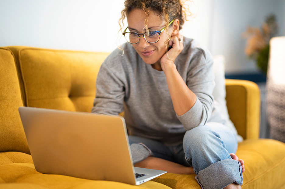 Woman on mustard yellow couch using her laptop to learn more about the Behavioral health program