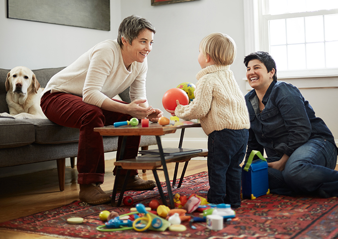 Male couple playing with their young son and toys in a living room, highlighting inclusive family care and benefits that support healthy households.
