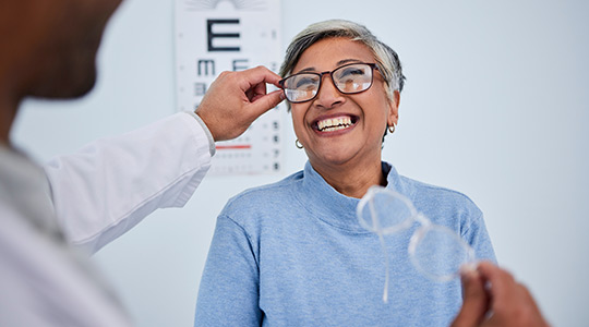 Woman with gray hair smiling as an eye doctor places glasses on her, highlighting how vision care helps support healthy aging.
