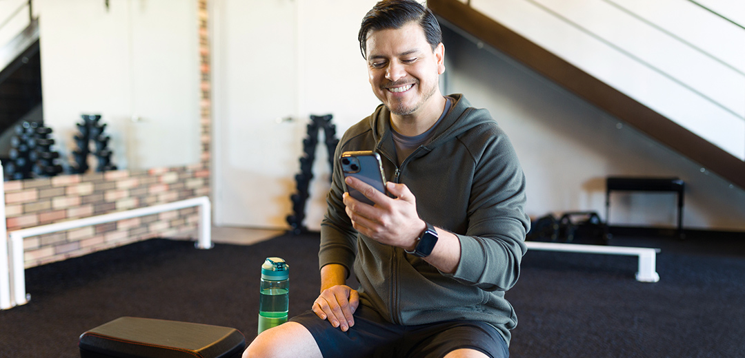 Smiling man uses Moda 360 health programs on his smartphone at home, supporting personalized wellness and healthier everyday choices.