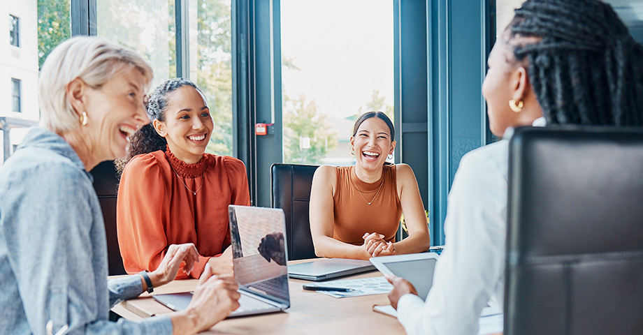 A group of professional women laugh together at a table, reflecting supportive connections and workplace well-being that contribute to overall health.
