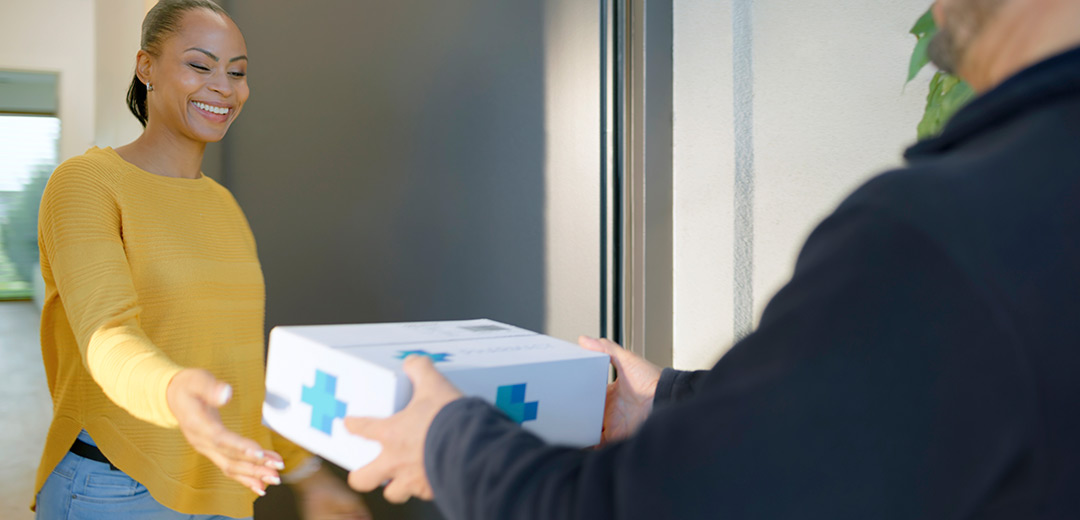 A smiling woman receives mail-order medications at her door, highlighting convenient access to prescriptions that supports timely and consistent care.