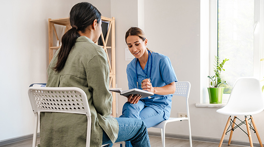 A medical assistant holding a pen and notebook asks a patient questions in a clean professional office setting.