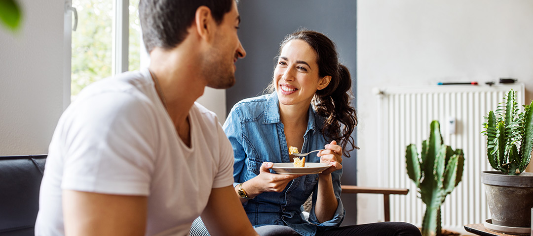 A couple smile at each other while eating together at home.