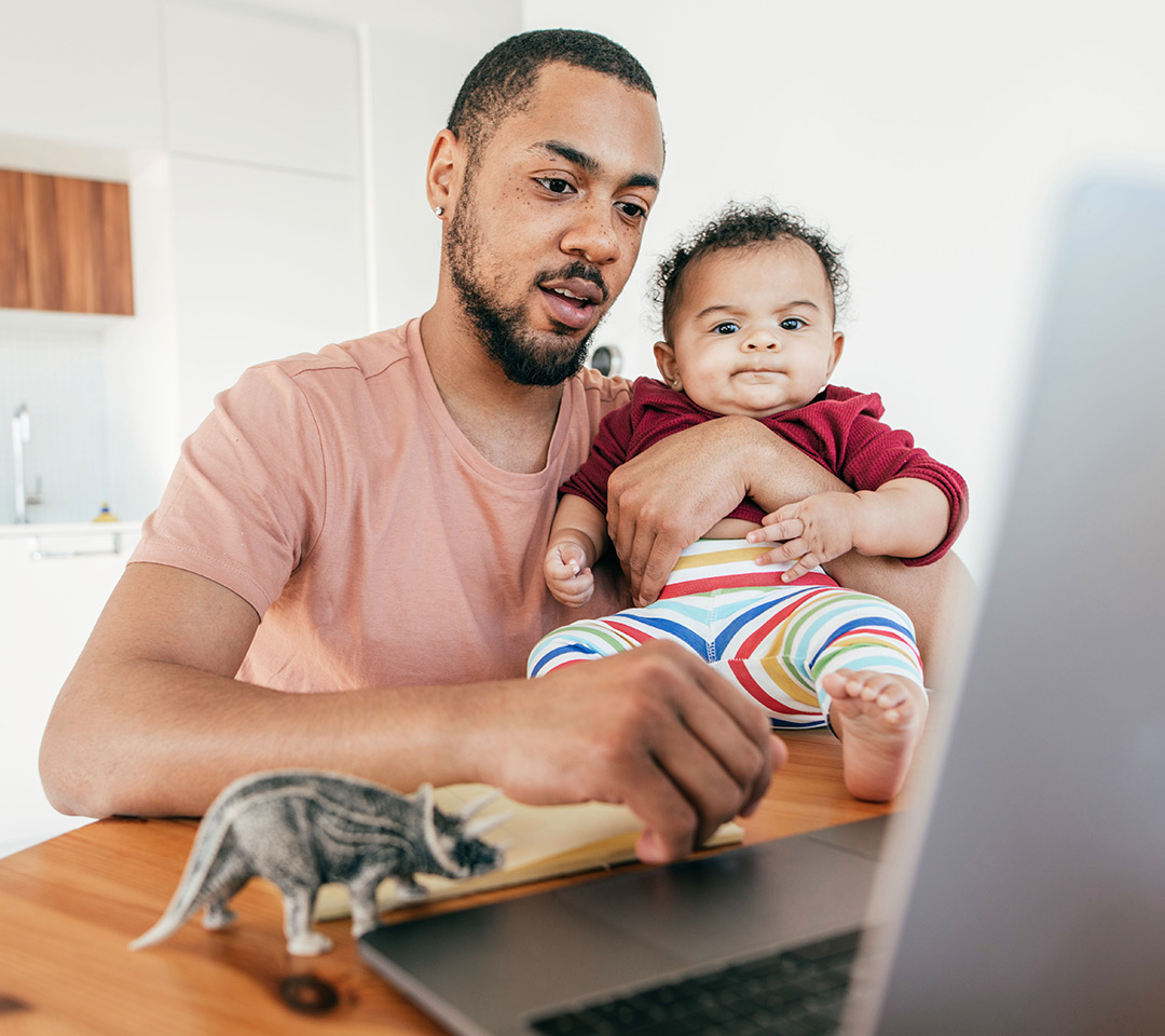 A young dad works on his laptop while holding his child.