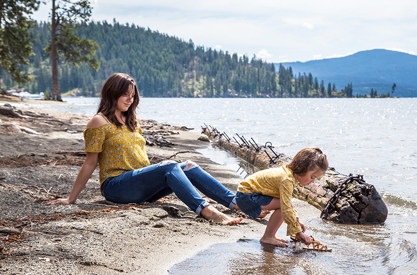 A mom sitting in the sand watching her daughter playing in the river 